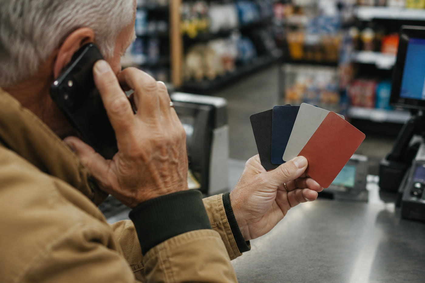 An older man at a store checkout, phone pressed to his ear, holding a fan of gift cards — the moment the scam wants